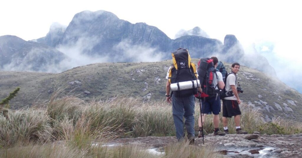 Hikers with backpacks trekking through Serra dos Órgãos, Petrópolis, with fog-covered mountain peaks in the background.