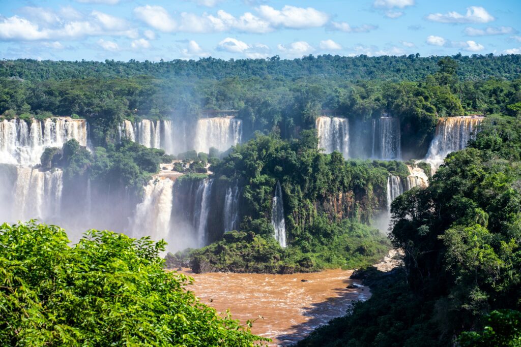 A panoramic view of Iguaçu Falls with multiple waterfalls cascading into a wide river, surrounded by lush green forest.