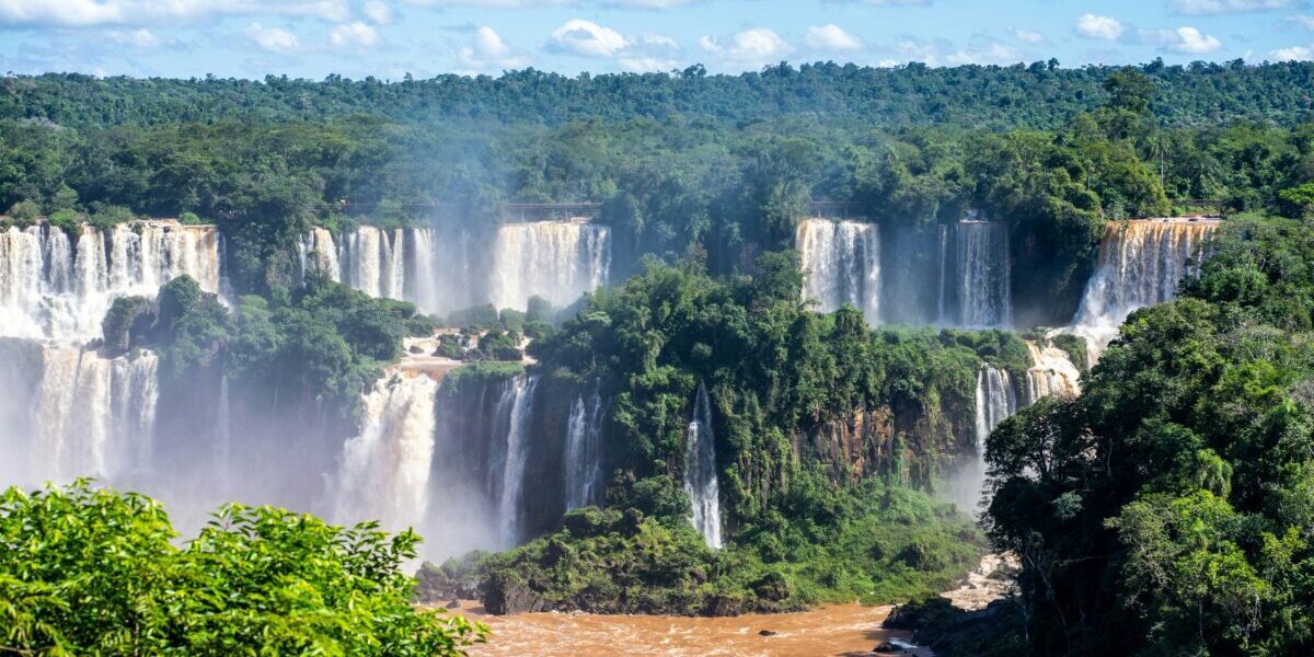 A panoramic view of Iguaçu Falls with multiple waterfalls cascading into a wide river, surrounded by lush green forest.