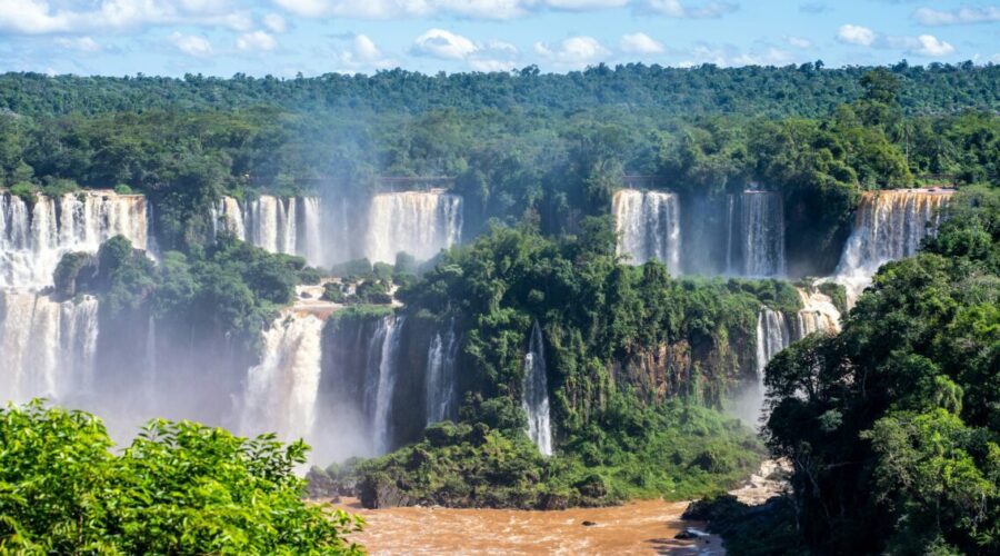 A panoramic view of Iguaçu Falls with multiple waterfalls cascading into a wide river, surrounded by lush green forest.