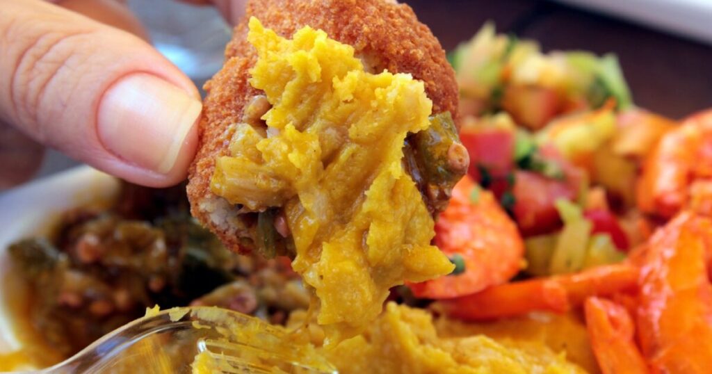 Person holding an acarajé filled with vatapá, with a plate of acarajé and shrimp in the background