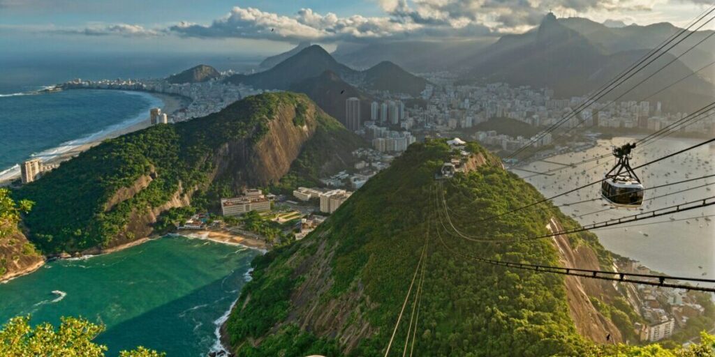 View from Sugarloaf Mountain showing the urban landscape of Rio de Janeiro, Christ the Redeemer partially obscured by clouds, and the ocean in the background.